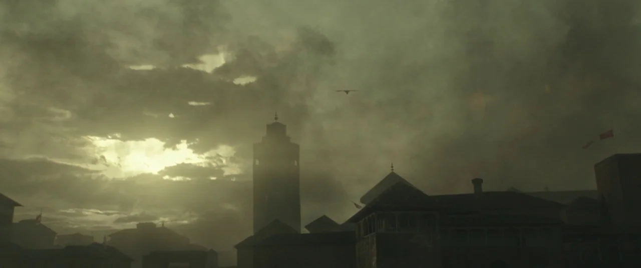 Assassin’s Creed (2016) movie still: establishing — An urban skyline features a central stone tower and rooftops under a heavy, cloudy sky. A…
