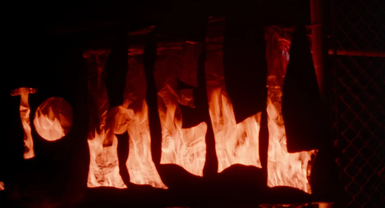 The Doom Generation (1995) movie still: closeup — A chain link fence at night features a stencil spelling the word TO HELL. Bright flames burn…