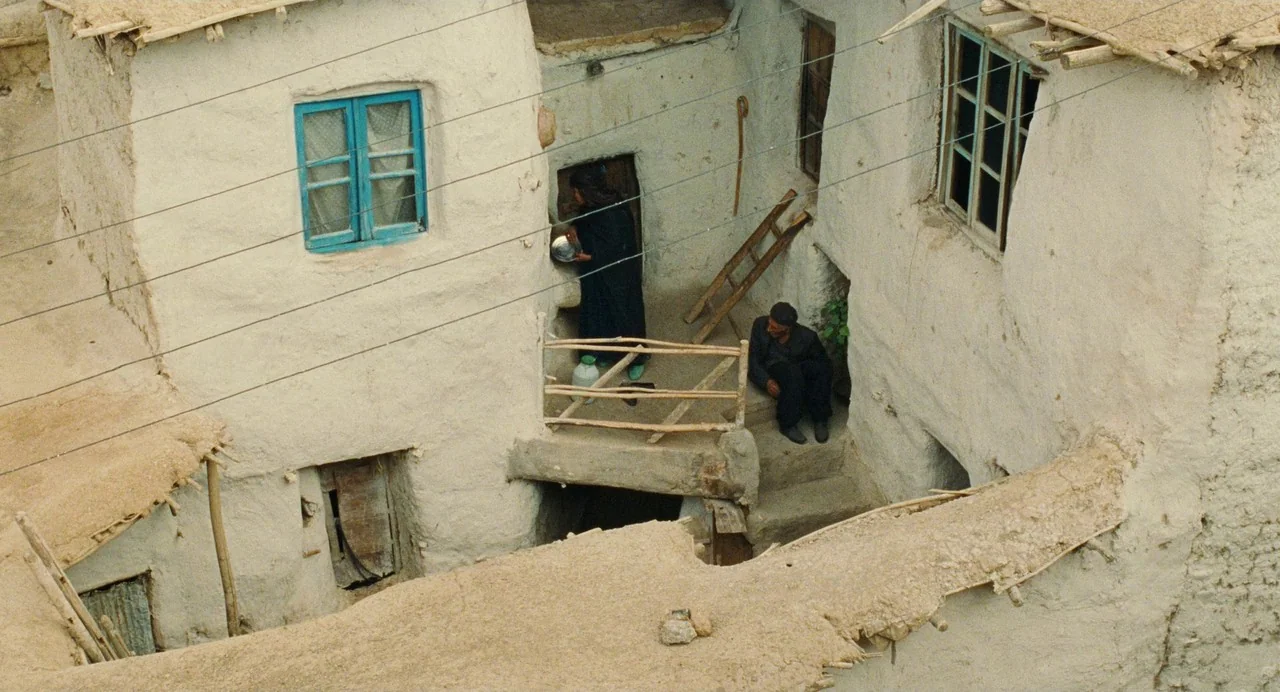 The Wind Will Carry Us (1999) movie still: wide — An austere adobe structure features white mud-plaster walls, prominent blue window frames, and a…