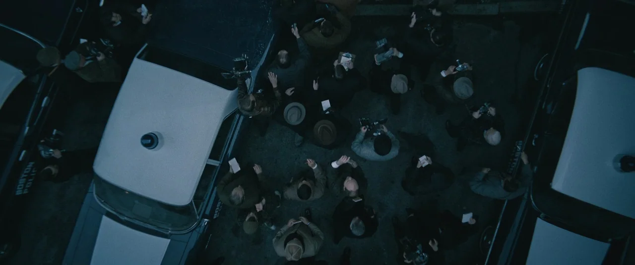Boston Strangler (2023) movie still: wide — A top-down view shows a crowd of people surrounding two police vehicles on asphalt. The individuals hold…