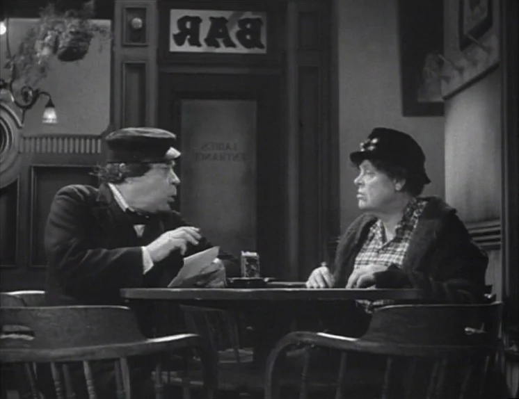 Anna Christie (1930) movie still: medium — A dimly lit tavern interior features a 'BAR' sign over a glass-paneled door. Two men sit at a round…