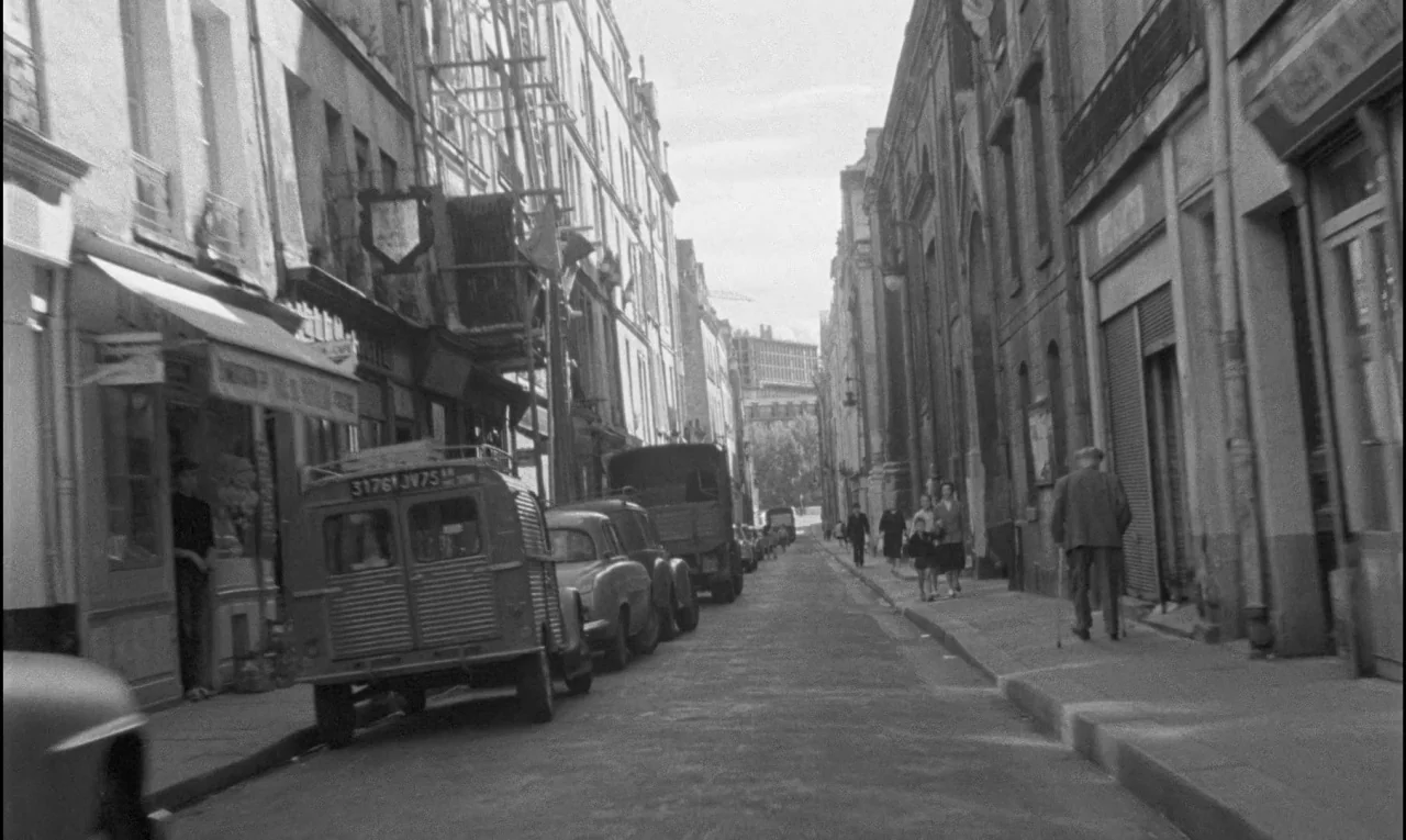 Bay of Angels (1963) movie still: establishing — A narrow European city street lined with multi-story buildings and parked vintage vehicles. Several…