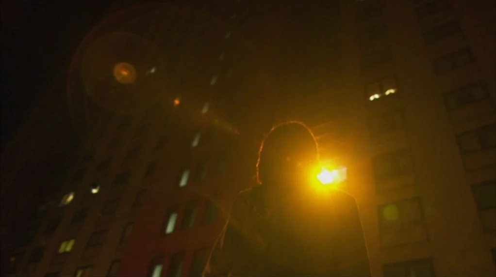 Red Road (2006) movie still: medium — A dark city street at night features a large building with illuminated windows above. A person stands facing…