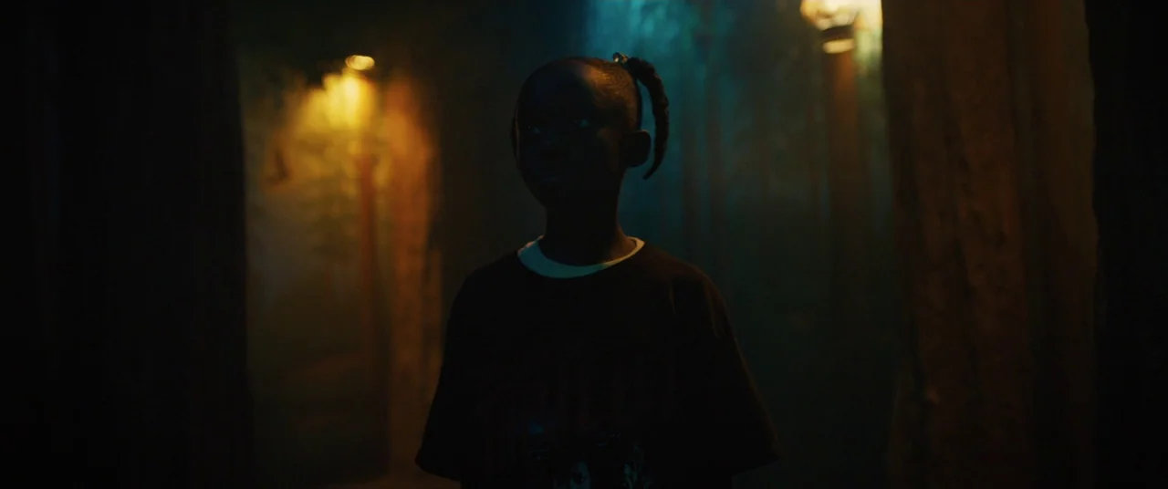 Us (2019) movie still: medium — A dark, wooded area features glowing lanterns hanging in the background trees. A young child stands centered in the…