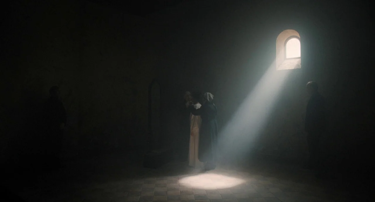 The First Omen (2024) movie still: wide — A dark stone room contains a single arched window letting in a bright beam. Two women stand within the…