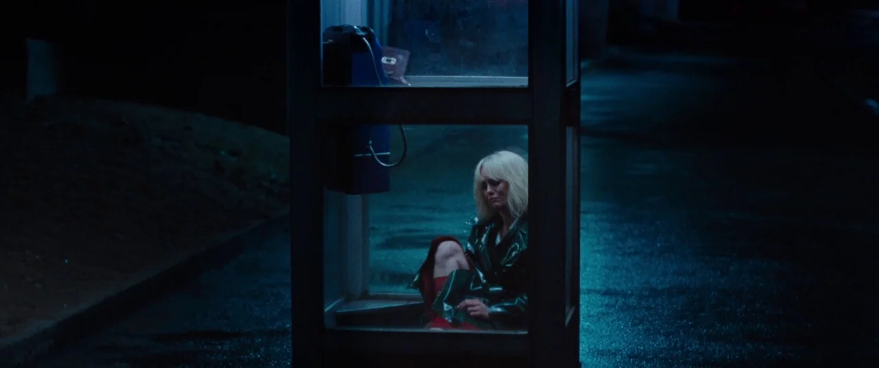 Knife+Heart (2018) movie still: wide — A glass phone booth stands on a dark, wet asphalt surface at night. A woman in a green coat sits on the floor…