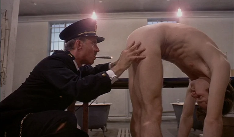 A Clockwork Orange (1971) movie still: medium — A utilitarian communal space with tiled walls and exposed ceiling light bulbs. A uniformed officer…