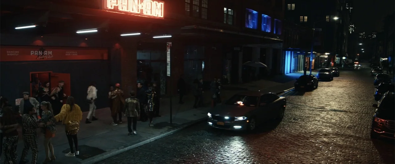 21 Bridges (2019) movie still: wide — A nighttime urban street with a nightclub entrance featuring a bright red neon sign. A group of people gathers…
