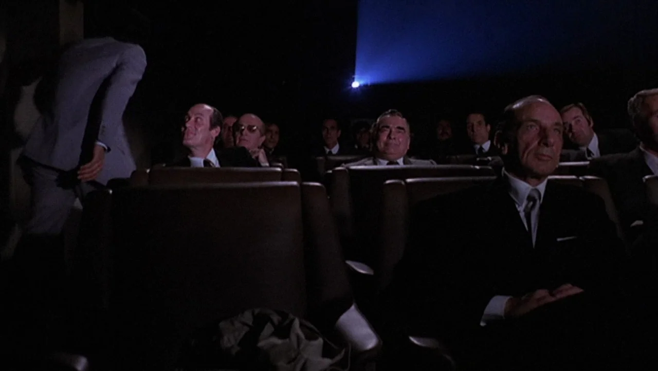 The Black Belly of the Tarantula (1971) movie still: wide — A dark movie theater features rows of padded seats occupied by men in suits. The room is…