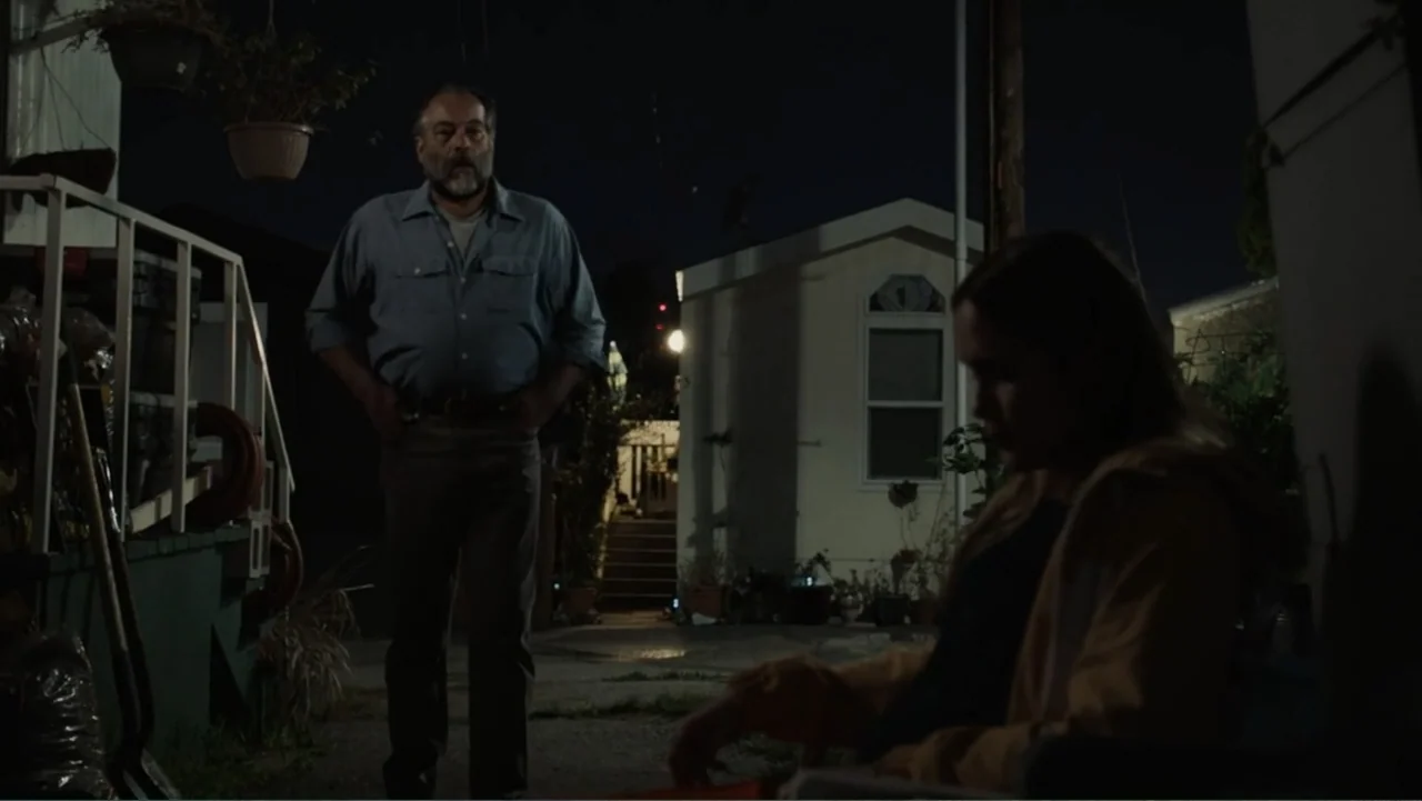 The Cleaner (2021) movie still: medium — An outdoor area at night with white mobile homes and hanging plants visible. An older man stands while…