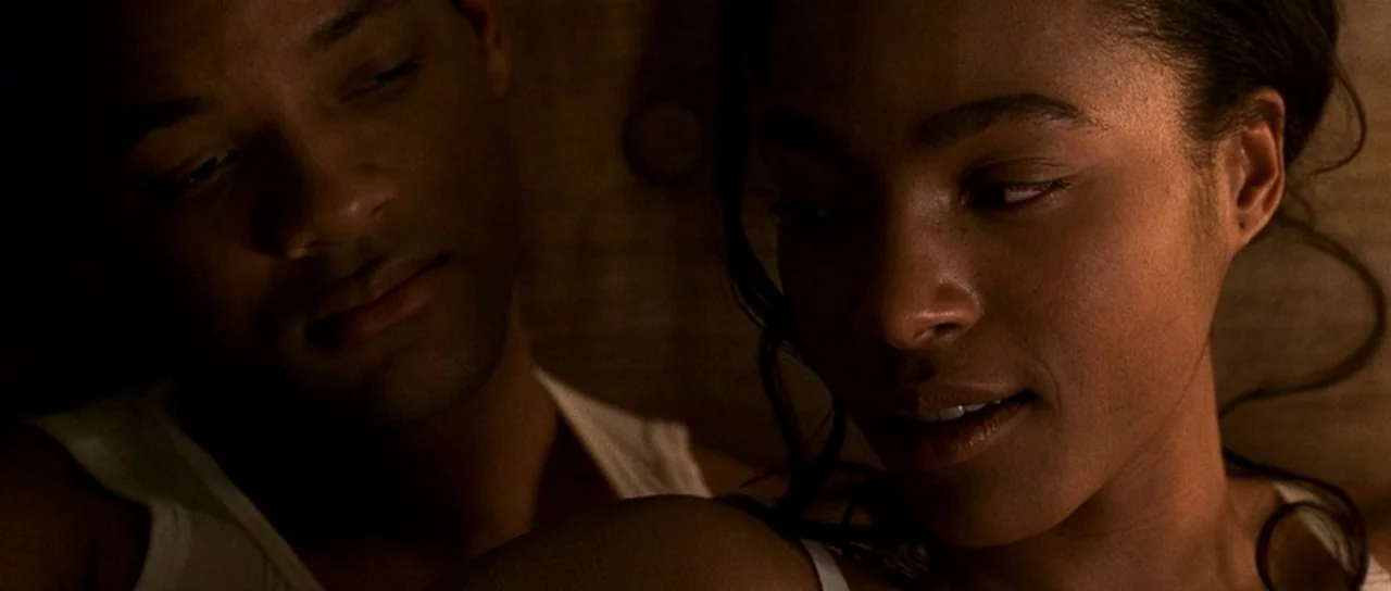 Ali (2001) movie still: closeup — A dimly lit room with dark wooden paneling in the background. A man and a woman lie close together in a low-light…