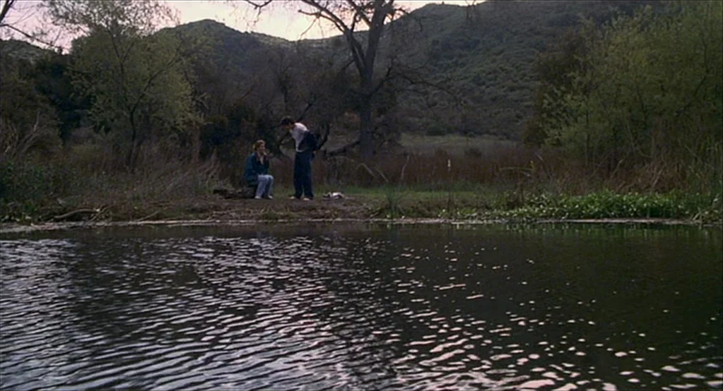 The Good Girl (2002) movie still: wide — A quiet pond bank bordered by dry reeds and low rolling hills. A woman sits on a small log while a man…