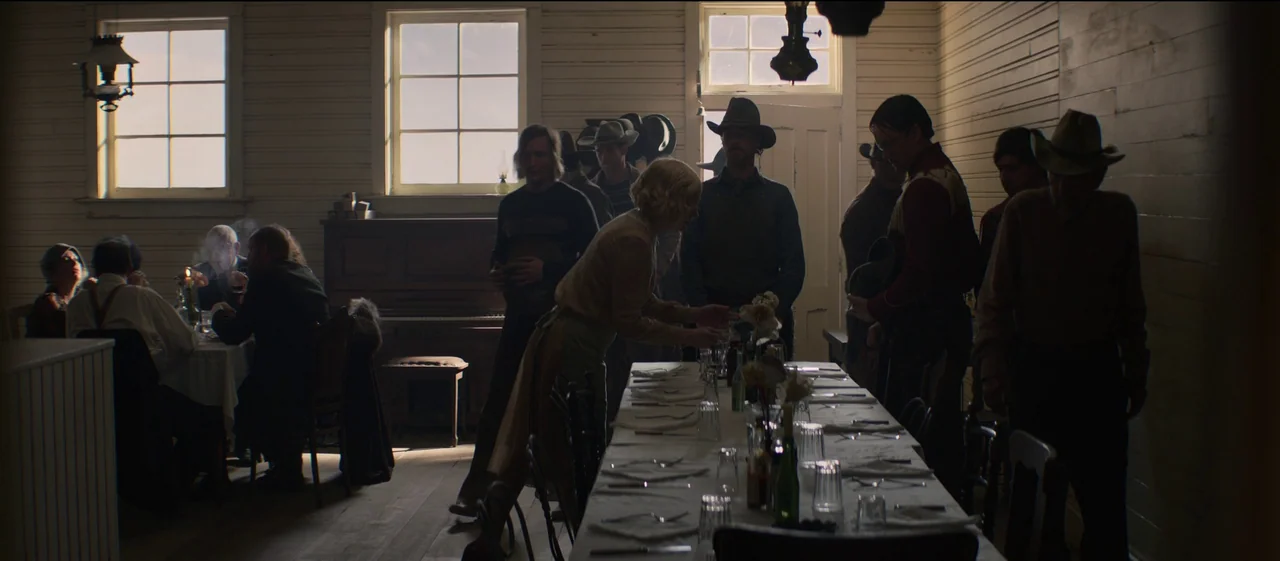 The Power of the Dog (2021) movie still: wide — A rustic wood-walled dining hall features long tables set for a communal meal. A woman organizes…