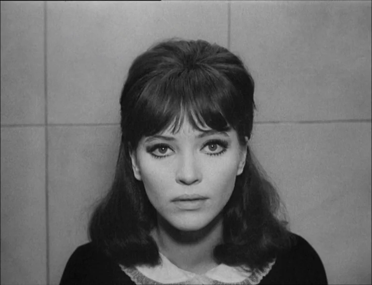Alphaville (1965) movie still: closeup — A woman stands centered in front of a flat, grid-patterned tiled surface. She stares directly forward into…