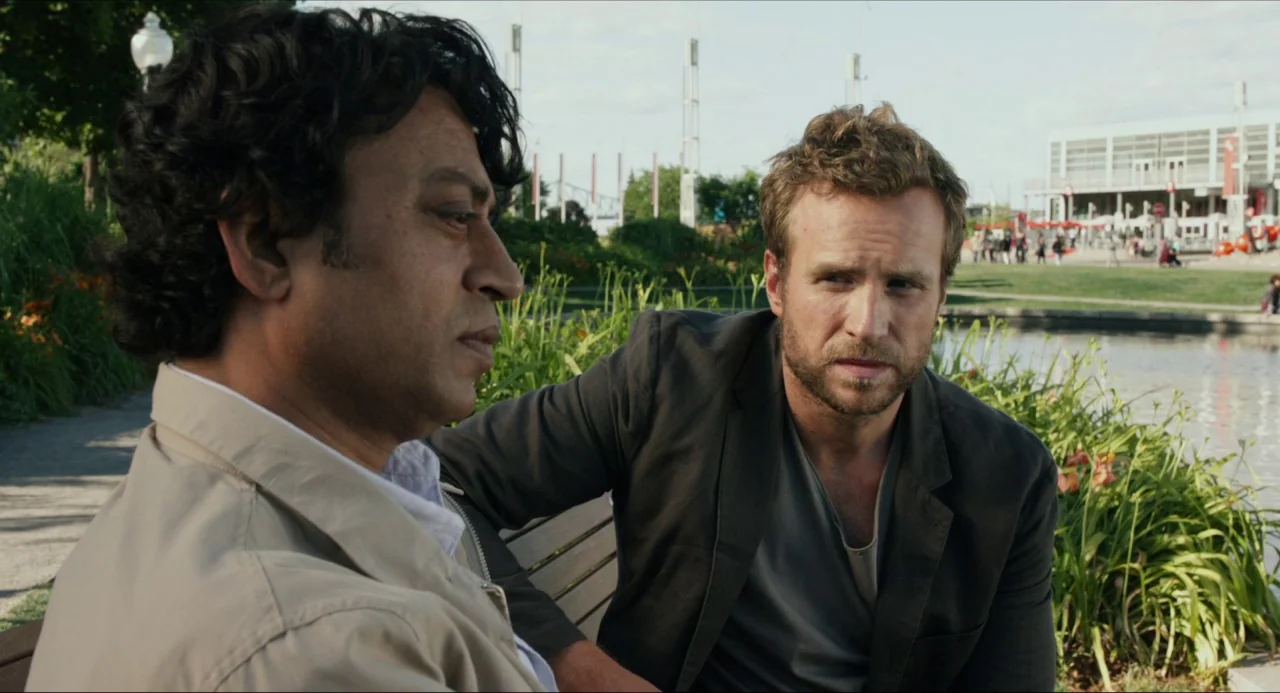 Life of Pi (2012) movie still: medium — An outdoor park setting with a small lake and a modern building in background. Two men sit together on a…