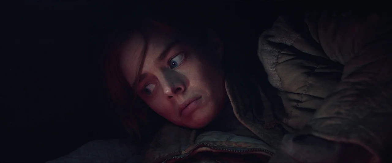 Azrael (2024) movie still: closeup — A person lies in a dark, enclosed environment with minimal ambient light visible. The subject turns their head…