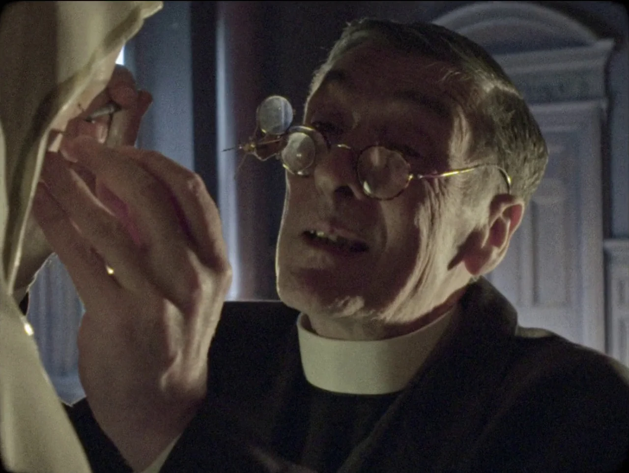 The Devil’s Doorway (2018) movie still: closeup — An elderly priest sits in a dimly lit study at night. He concentrates while examining a small…
