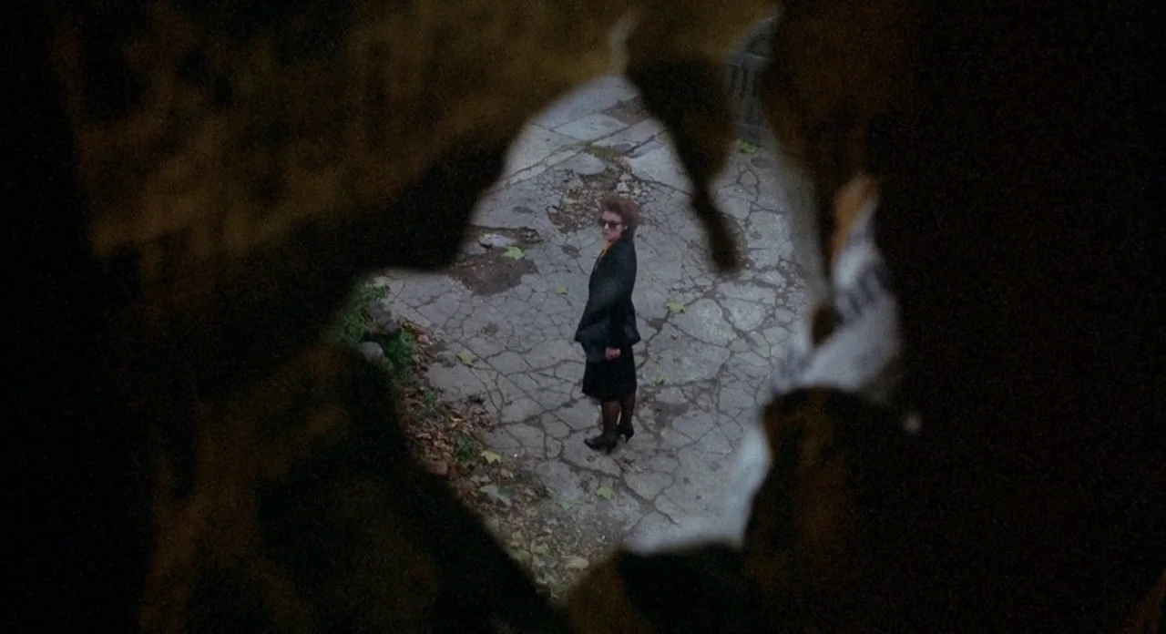 Hellraiser (1987) movie still: wide — A woman stands in a desolate, cracked concrete courtyard viewed through a dark frame. She pauses on the…