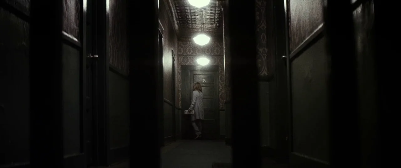 Lords Of Salem (2013) movie still: wide — A dimly lit narrow corridor features patterned wallpaper and multiple dark doors. A woman stands in front…