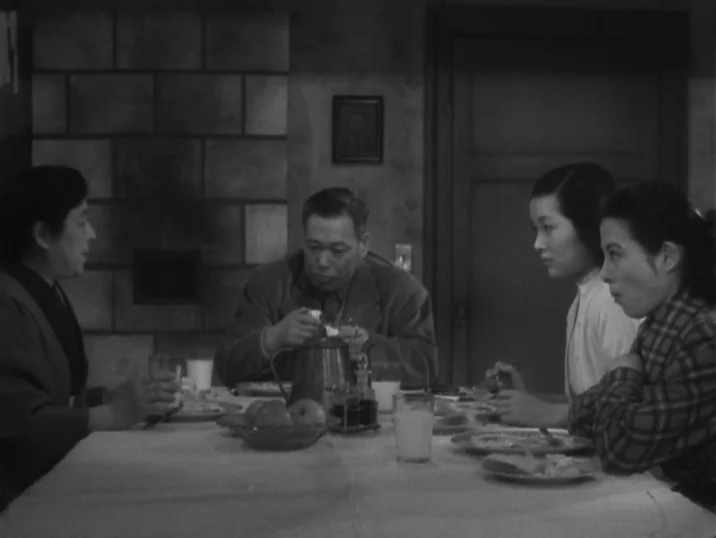 The Idiot (1951) movie still: medium — A domestic dining space with a tiled fireplace feature in the rear. Four people are seated at a table eating…