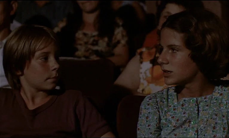 Small Change (1976) movie still: medium — A dimly lit movie theater with rows of seats occupied by children. A boy and a girl are seated next to…