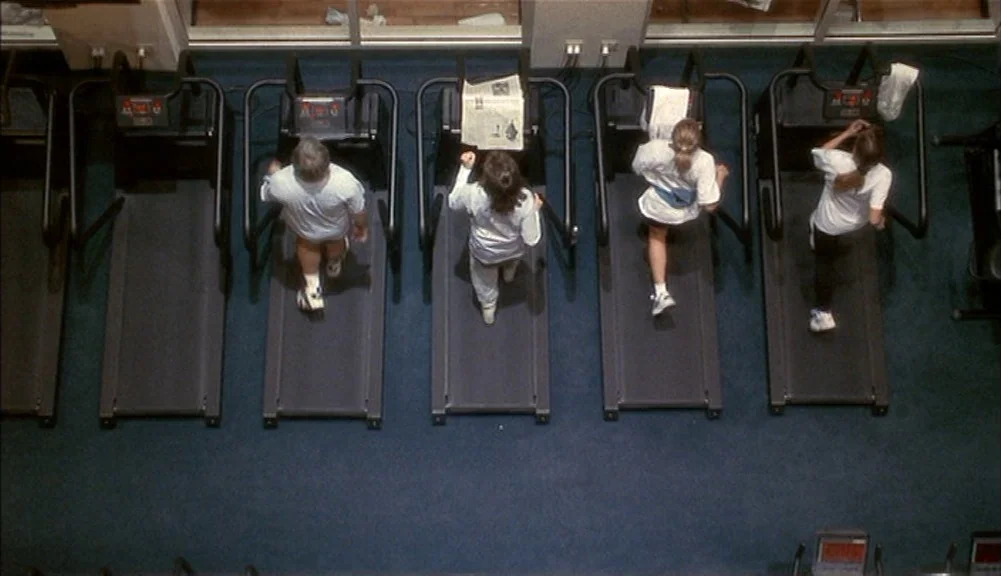 The Mirror Has Two Faces (1996) movie still: wide — A row of four people run on treadmills in a brightly lit indoor space. The high-angle view looks…