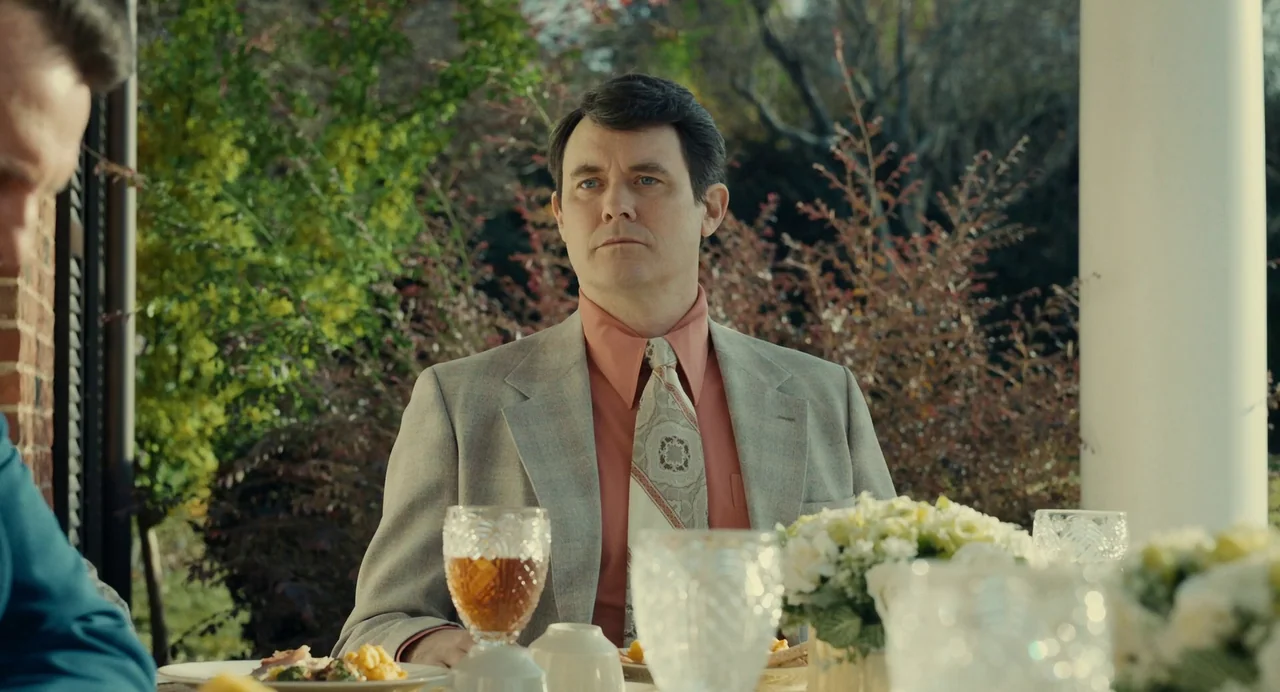 The Eyes of Tammy Faye (2021) movie still: medium — A man in a 1970s suit sits at a table on a patio. The background consists of out-of-focus trees…