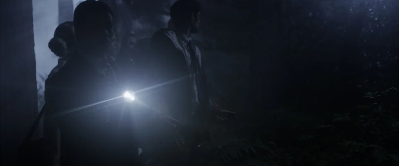 Howl (2015) movie still: medium — A dark night setting outdoors among dense vegetation and shadows. Two people stand close together using a…
