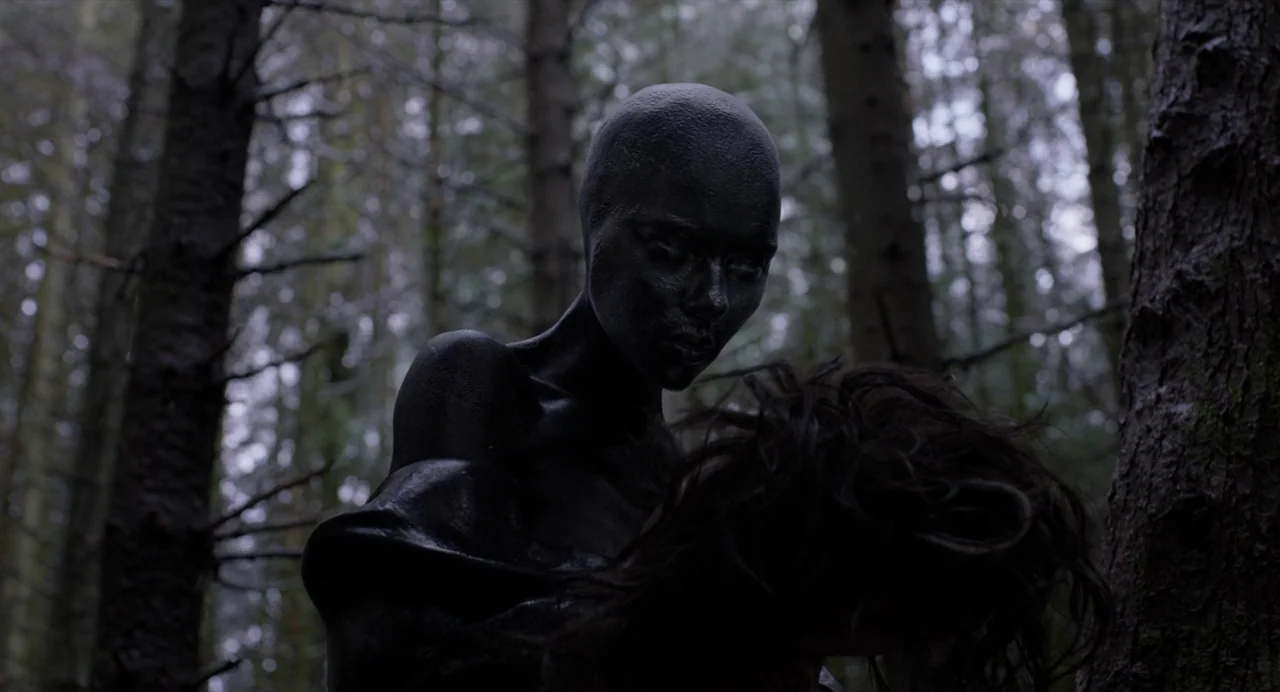 Under The Skin (2013) movie still: medium — A dense cluster of tall pine trees forms the backdrop of the scene. A bald individual with skin coated…
