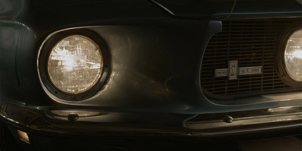 The Pickup (2025) movie still: closeup — A close view shows the front grille and headlight of a classic vintage car. The illuminated round headlamp…