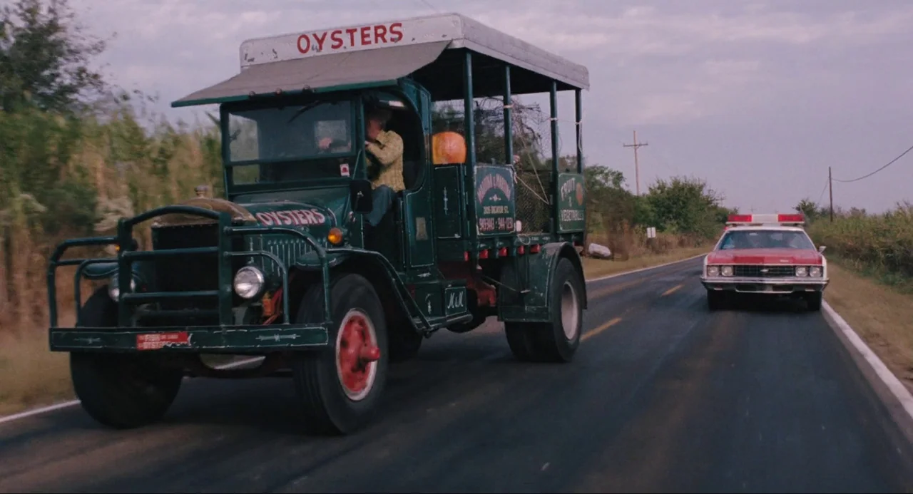 Live and Let Die (1973) movie still: wide — A green open-air truck labeled OYSTERS drives down an asphalt road during daytime. A red and white…
