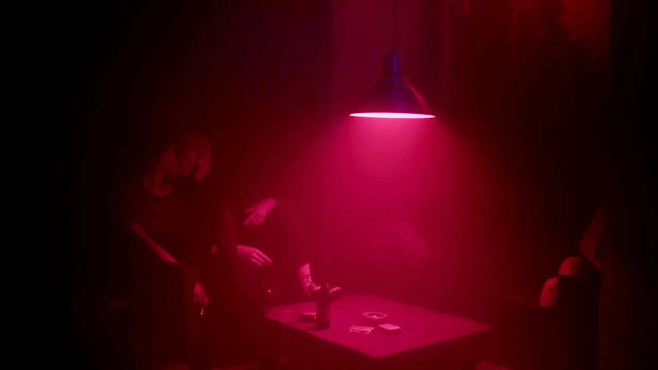 The Last Year of Darkness (2023) movie still: medium — A dark space lit primarily by a single overhead pendant light emitting red illumination. A…
