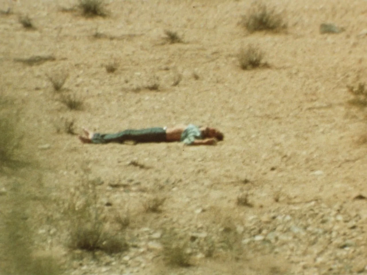 Punishment Park (1971) movie still: wide — An arid desert landscape features sparse dry brush on a coarse sandy surface. A solitary person lies…