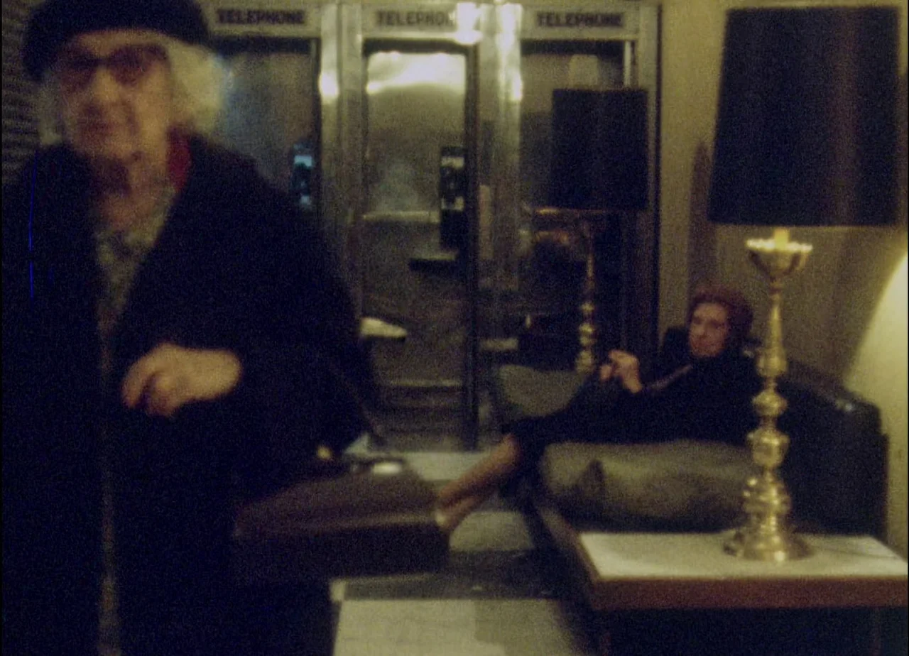 Hotel Monterey (1973) movie still: medium — A dimly lit indoor lobby features a series of telephone booths in the back. An elderly woman stands in…