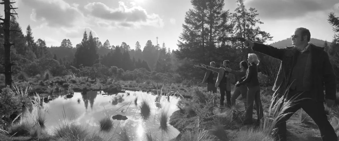 Roma (2018) movie still: wide — A group of four people line up beside a pond in a wooded area. They aim handguns toward an unseen point while…