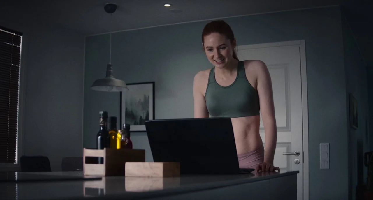 Dual (2022) movie still: medium — A modern space with a clean countertop and grey walls during the night. A woman in athletic wear stands at the…