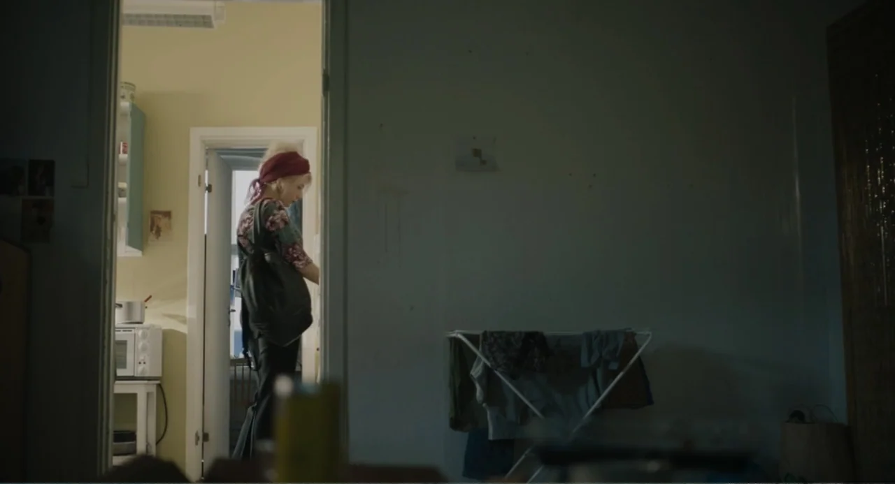 Ninjababy (2021) movie still: medium — A woman stands in the doorway of a simple room with pale yellow walls. She reaches toward an area inside the…