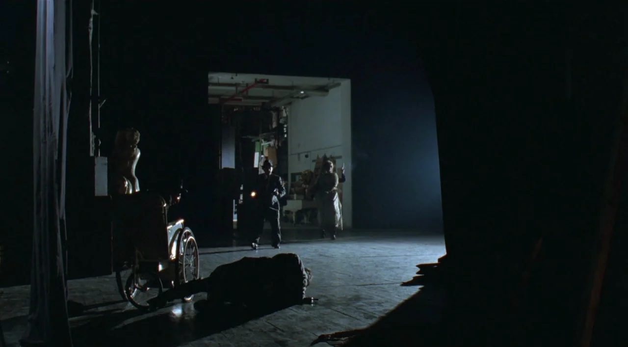 Underground (1995) movie still: wide — A dark wooden stage features a wheelchair on the left and a brightly lit doorway. A figure in a trench coat…