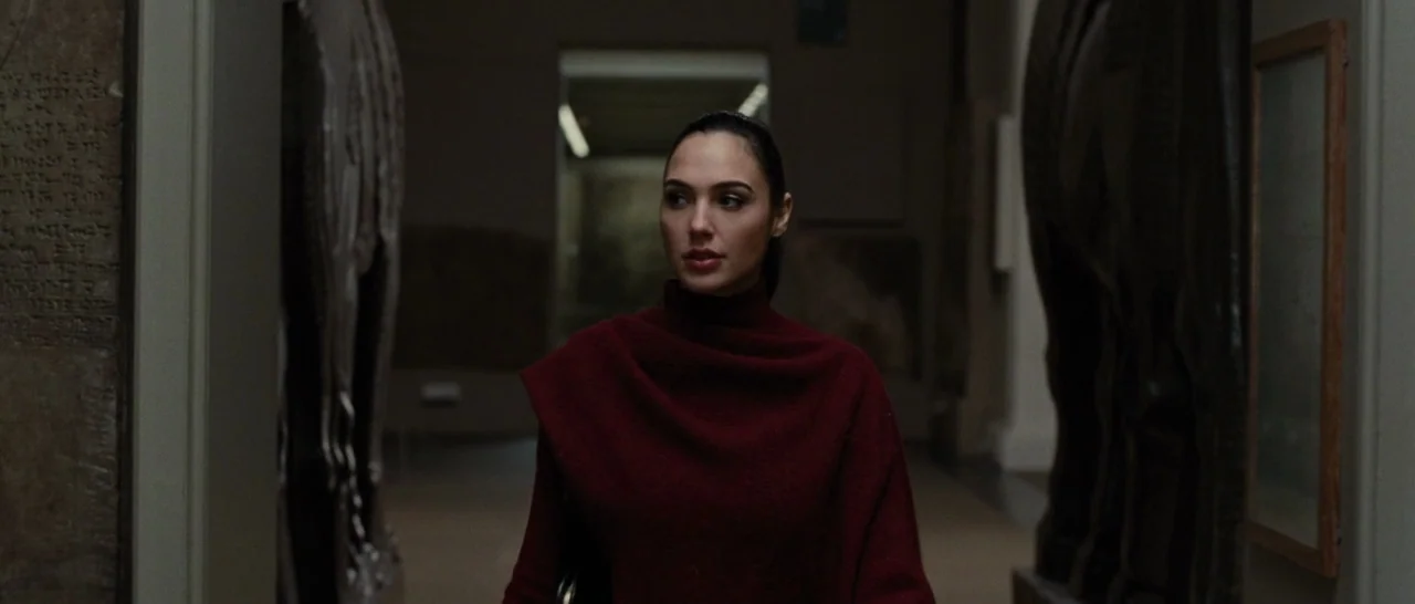 Wonder Woman (2017) movie still: medium — A dimly lit museum hallway flanked by large carved stone statues and inscribed tablets. A woman in a…