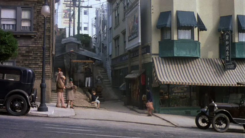 Hammett (1982) movie still: wide — A 1940s urban street corner features a Sun Wee Laundry shop with striped awnings. Several pedestrians stand near…