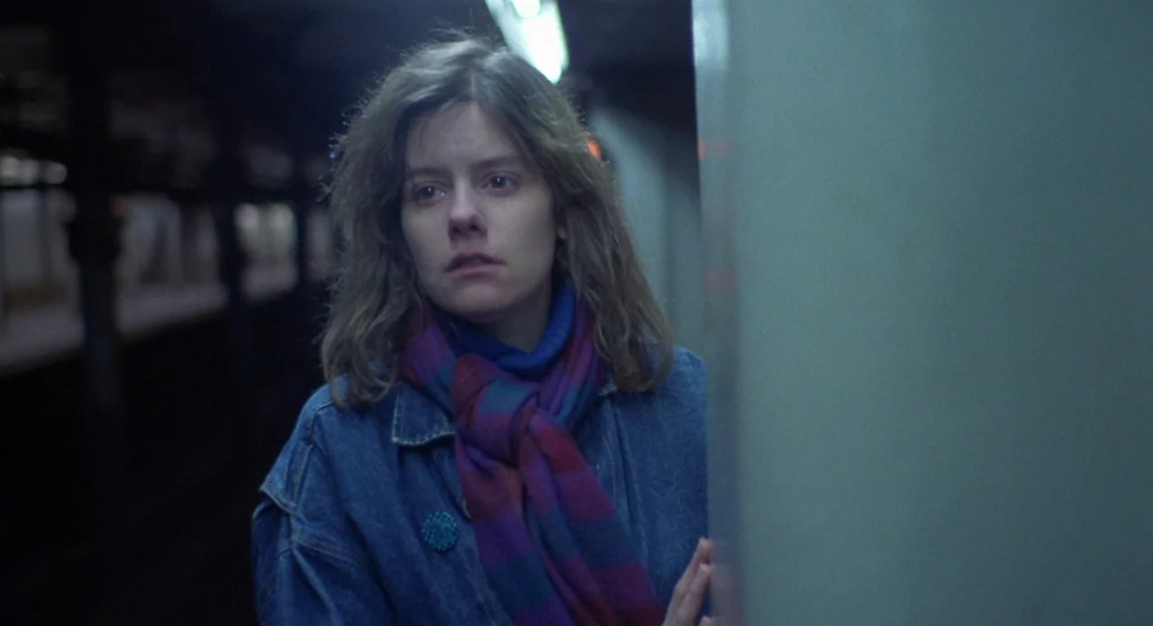 Brain Damage (1988) movie still: medium — A dimly lit subway station platform with metal structural columns and obscured tracks. A young woman…