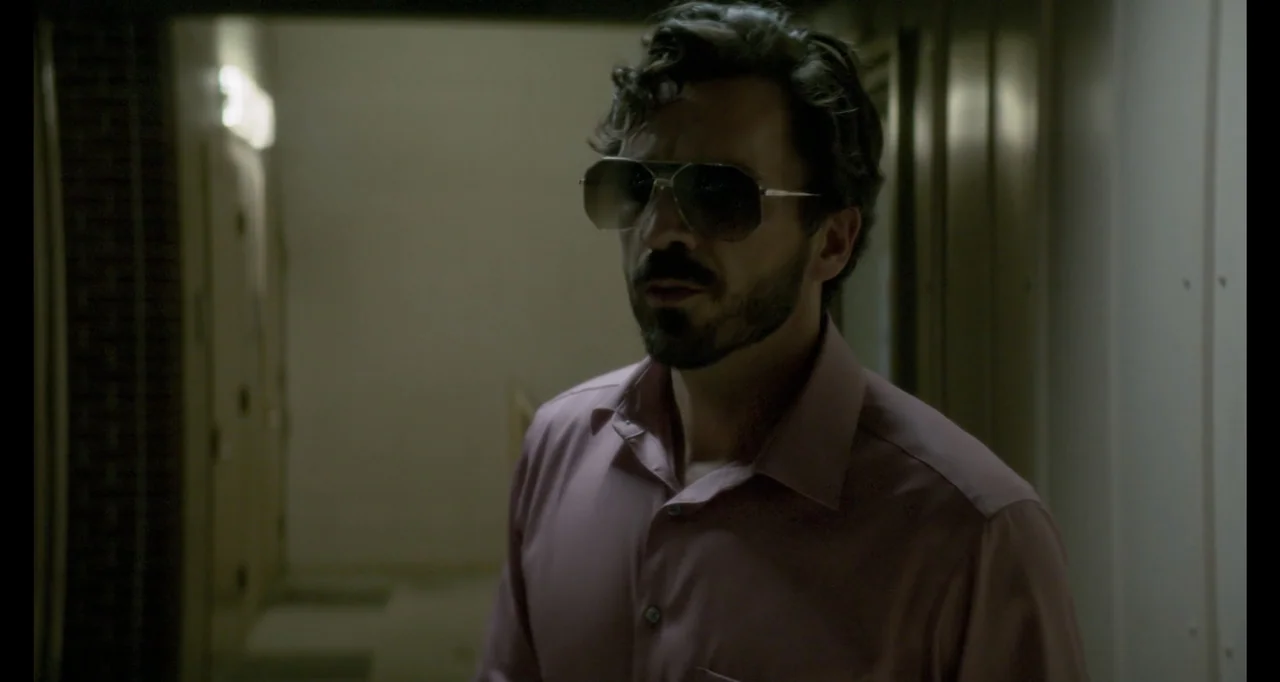 The Poison Rose (2019) movie still: medium — A dimly lit interior corridor features plain walls and muted tones. A man wearing tinted sunglasses…