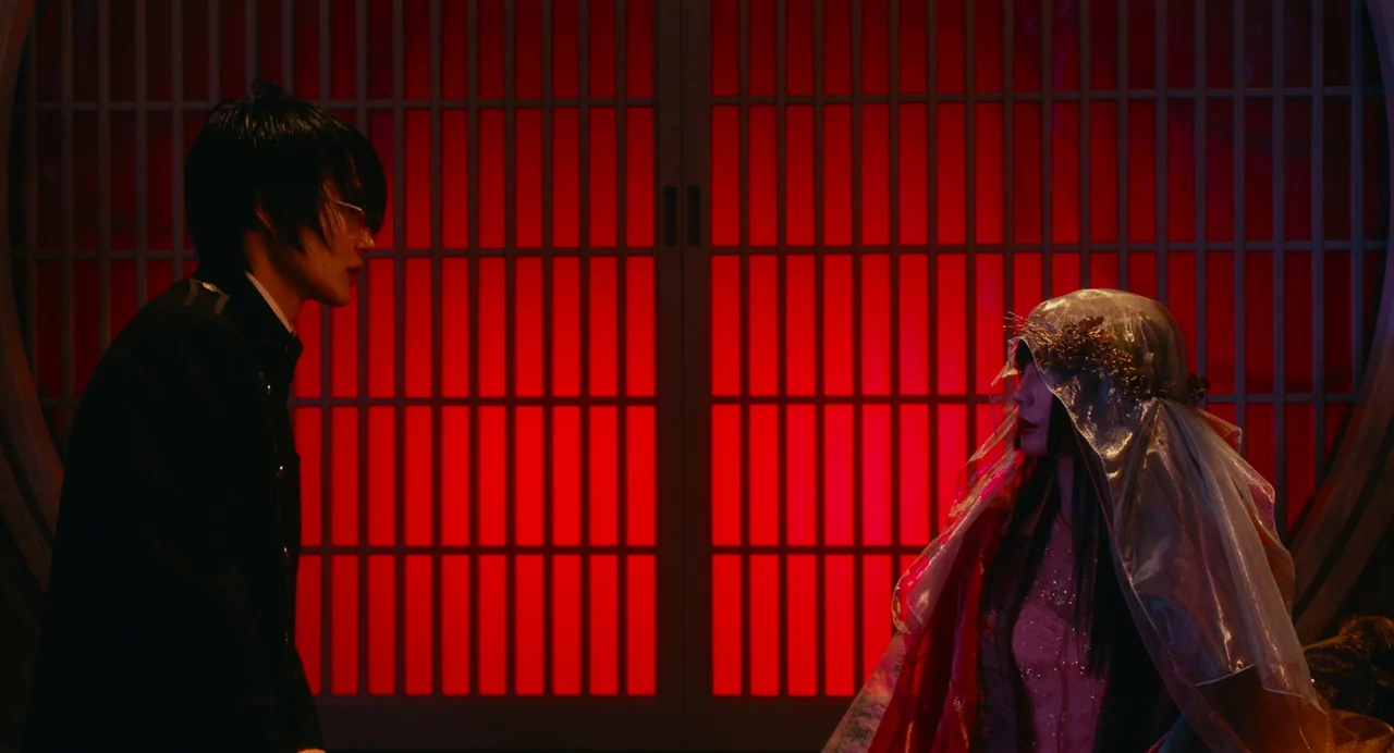 xxxHolic (2022) movie still: medium — A traditional Japanese room features a glowing red shoji screen in the background. A man in a dark coat faces…