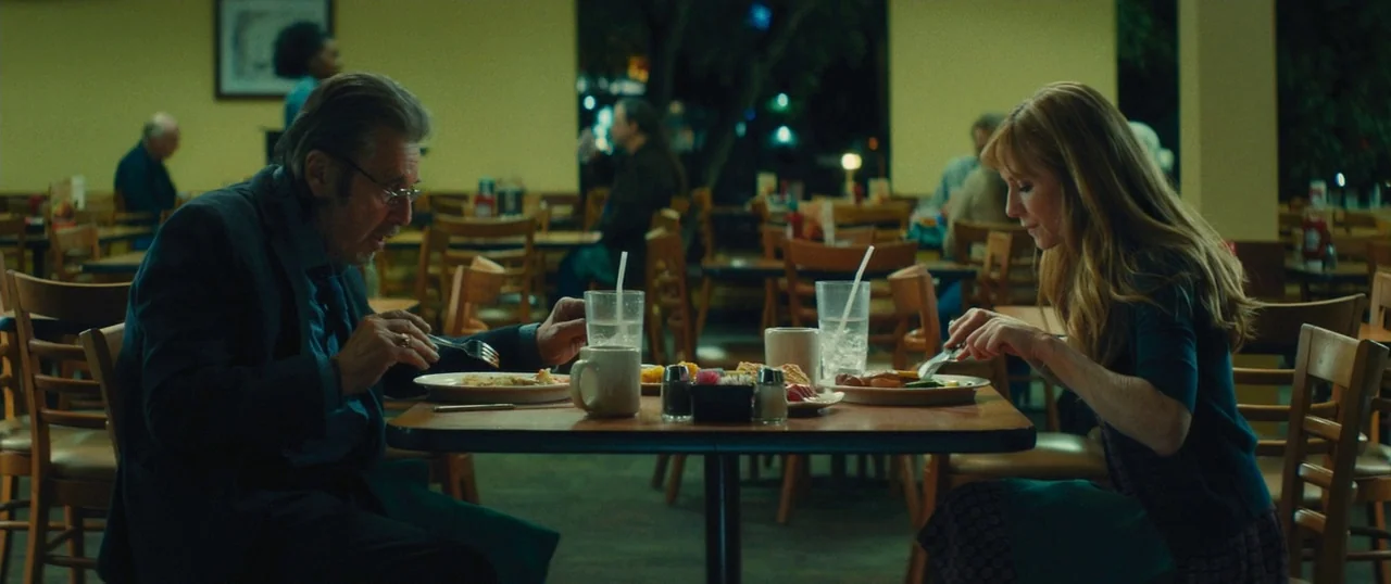 Manglehorn (2014) movie still: medium — A quiet diner interior with rows of wooden chairs and tables in low light. A man and woman eat their meals…