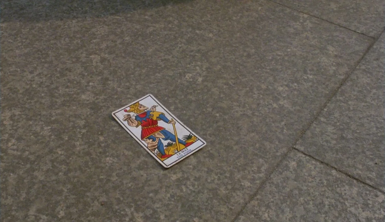 The Rainbow Thief (1990) movie still: closeup — A single tarot card rests on a grey concrete tiled ground. The card depicts the fool character from…