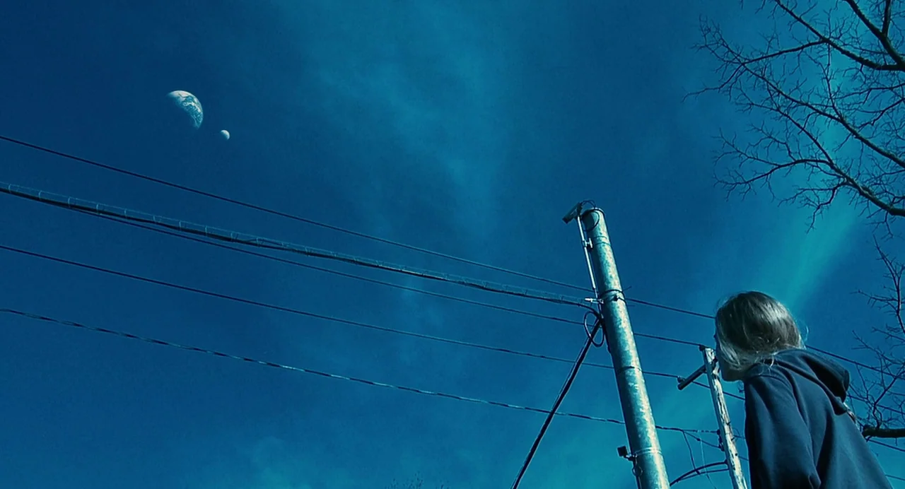 Another Earth (2011) movie still: wide — A dark blue twilight sky features the planet Earth and a smaller moon floating. A young woman in a hoodie…