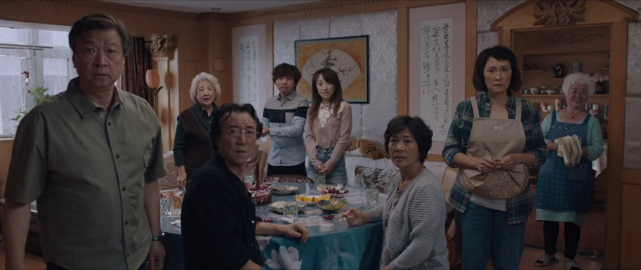 The Farewell (2019) movie still: fullbody — A residential room features a circular table surrounded by seven Asian individuals. They stand and sit…