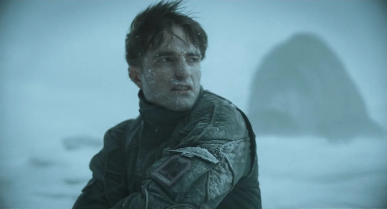 Mickey 17 (2025) movie still: medium — A man stands on a frozen shore with a large rock formation in the distance. The man is covered in snow and…