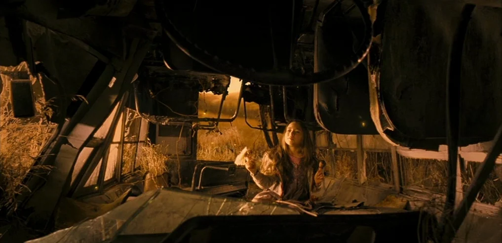 Tideland (2005) movie still: medium — The interior of a decaying, rusted bus is overgrown with tall dried grass. A young girl sits calmly among the…