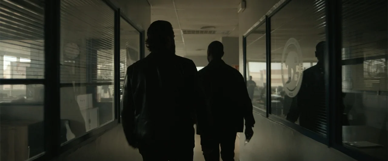 Den of Thieves 2: Pantera (2025) movie still: fullbody — A long interior office hallway features glass partitions with window blinds and white…